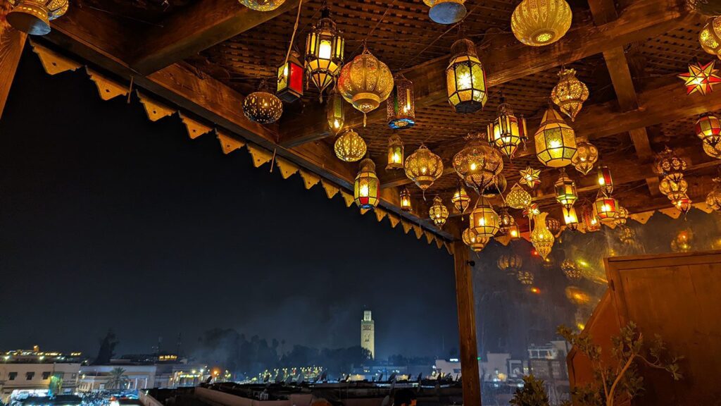 Romantic colorful hanging lanterns decorate a rooftop at night, overlooking a cityscape with a lit mosque tower in the distance.