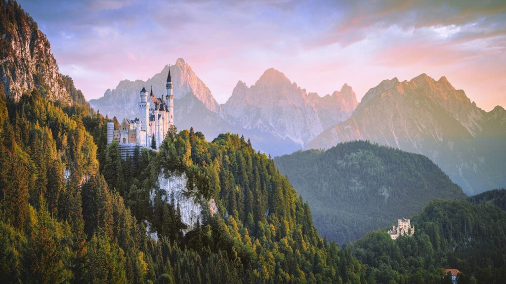 A panoramic view of Neuschwanstein Castle with surrounding forests and mountain backdrop at sunset