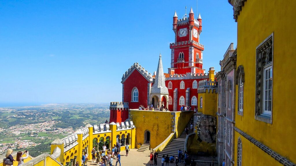 View of Pena Palace in Sintra, Portugal, showing its vibrant red and yellow towers and walls with tourists walking around on a sunny day, sure to inspire kids to travel.