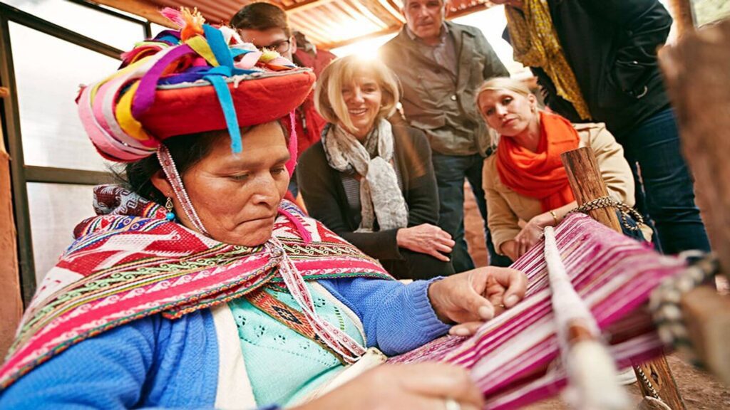 A woman in traditional Andean clothing demonstrates weaving techniques to an attentive group of tourists on a local culture tour.