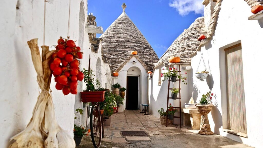 A traditional trullo house in Alberobello, Italy, ideal for family travel destinations, with a white stone facade, conical roofs, and decorations including hanging garlic and tomatoes.