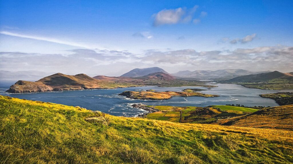 Panoramic view of a verdant Irish coastal landscape with rolling hills, a serene bay, and scattered islands under a vast blue sky in 2023.