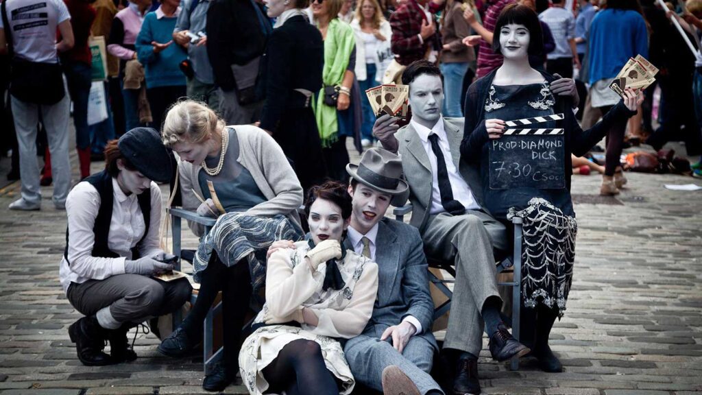 A group of five street performers dressed in vintage clothing, posing stoically as silent film actors on a bucket list event, with black and white makeup, amidst a crowd.