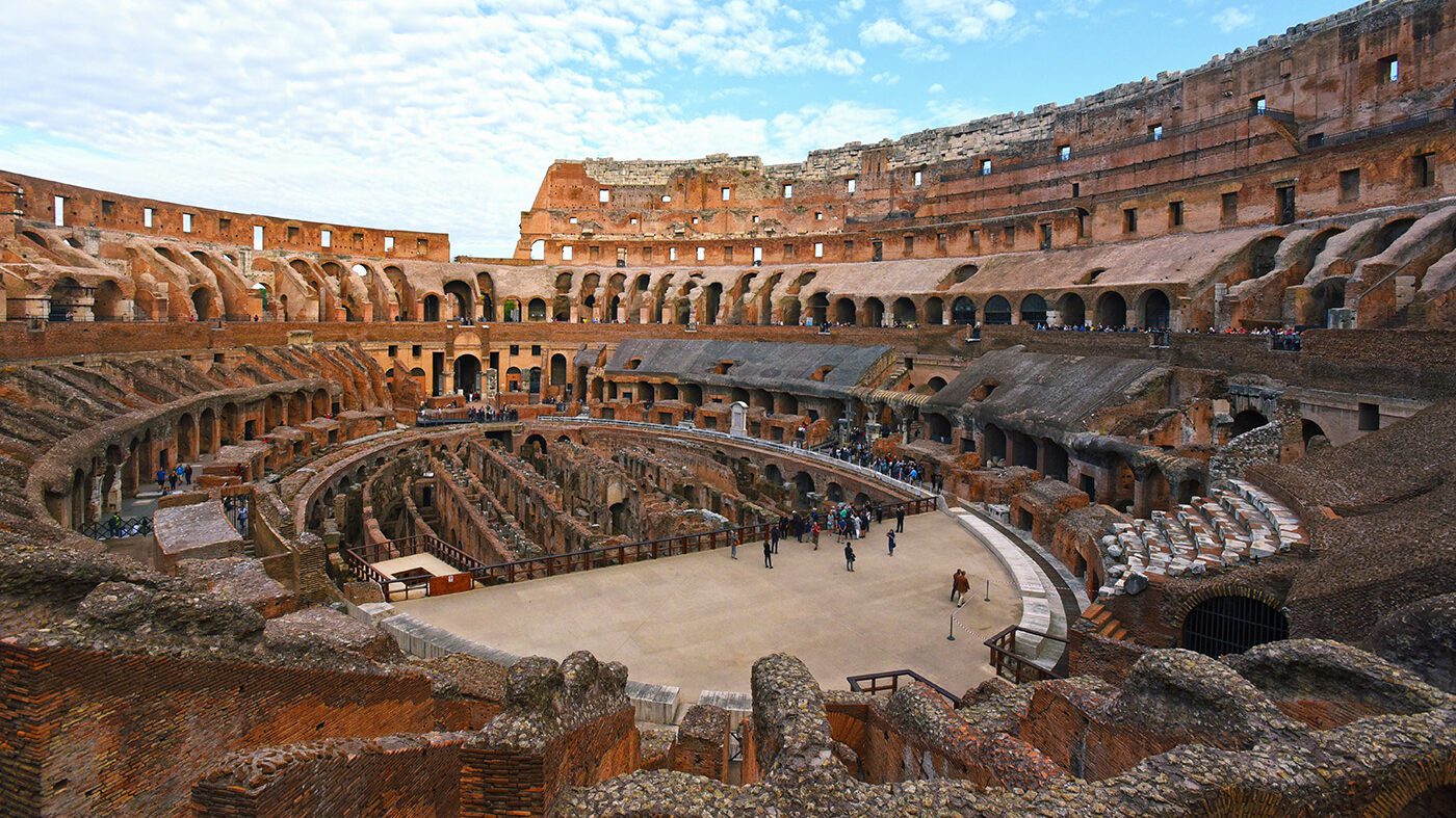 Interior view of the Colosseum in Rome, showing the arena, exposed underground structures, and tiers of arch-lined seating under a clear sky, a must-see on any European summer bucket list