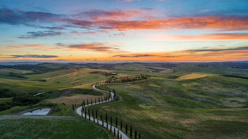 Aerial view of a winding road through rolling hills at sunset, with vibrant clouds above and patches of green and yellow fields below, capturing the essence of an Italy tour.