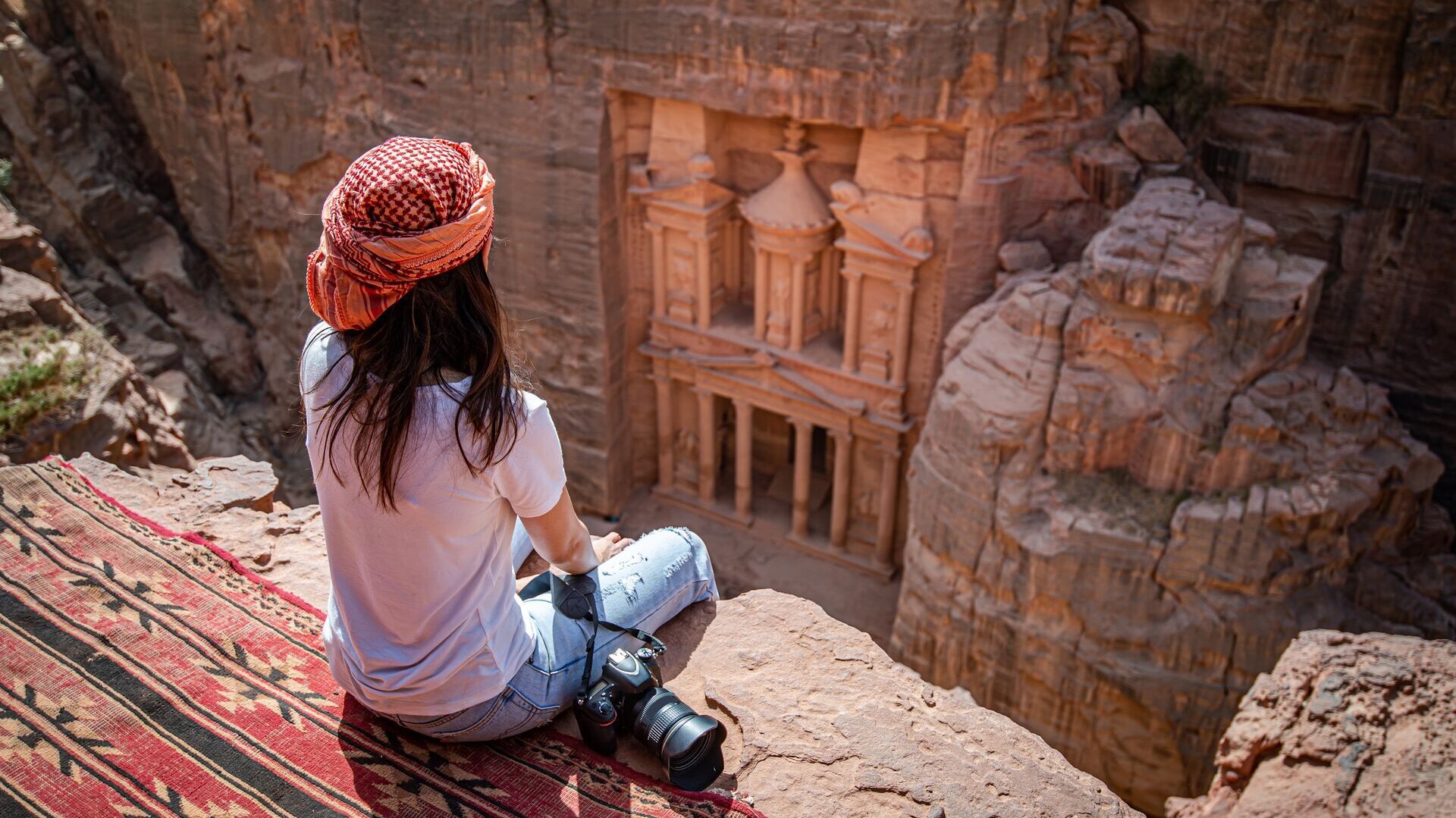 Woman in a head scarf sits atop a rock face overlooking the Treasury in Petra, Jordan
