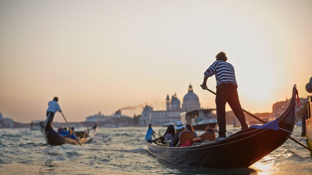 Gondoliers transporting guests in Venice through the canals, gondoliers are stood at the back of the boat, wearing striped black and white t-shirts.