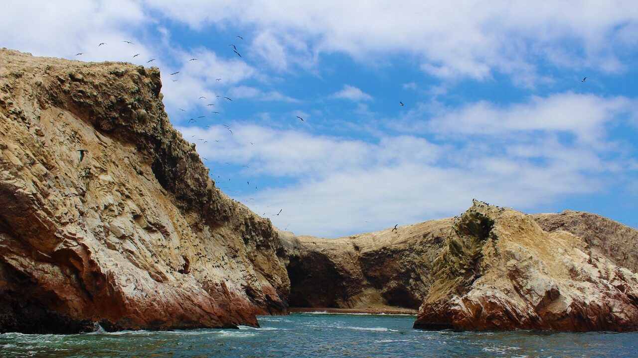 Rocky coastal cliffs in Peru with birds flying under a blue sky with clouds.