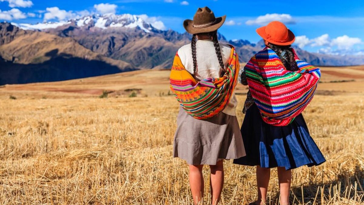 Two women in traditional Andean clothing and hats stand in a field looking at distant mountains while travelling responsibly.