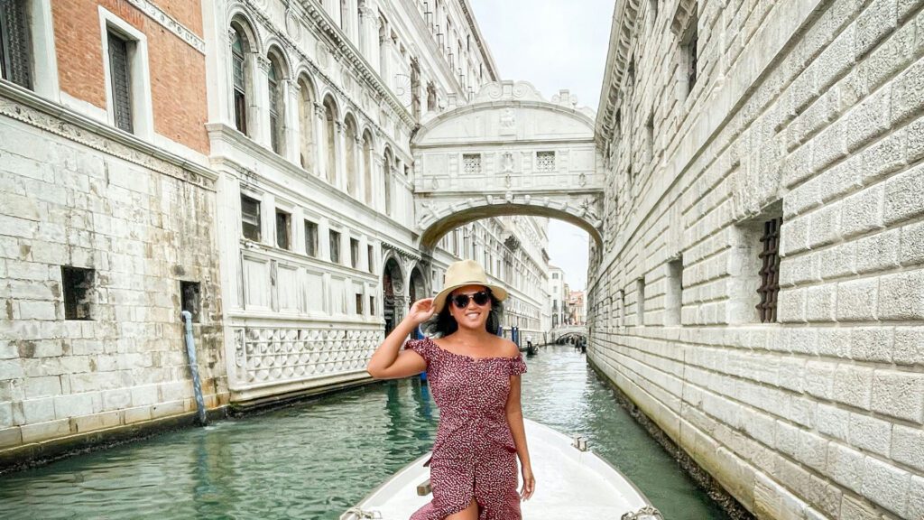 Woman wearing a straw hat and a red floral dress standing in a boat with Venice's Bridge of Sighs in the background