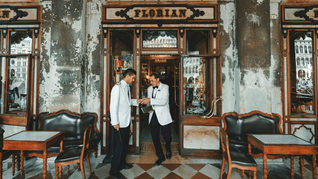 Two waiters dressed in traditional white jackets interact outside a historic looking cafe