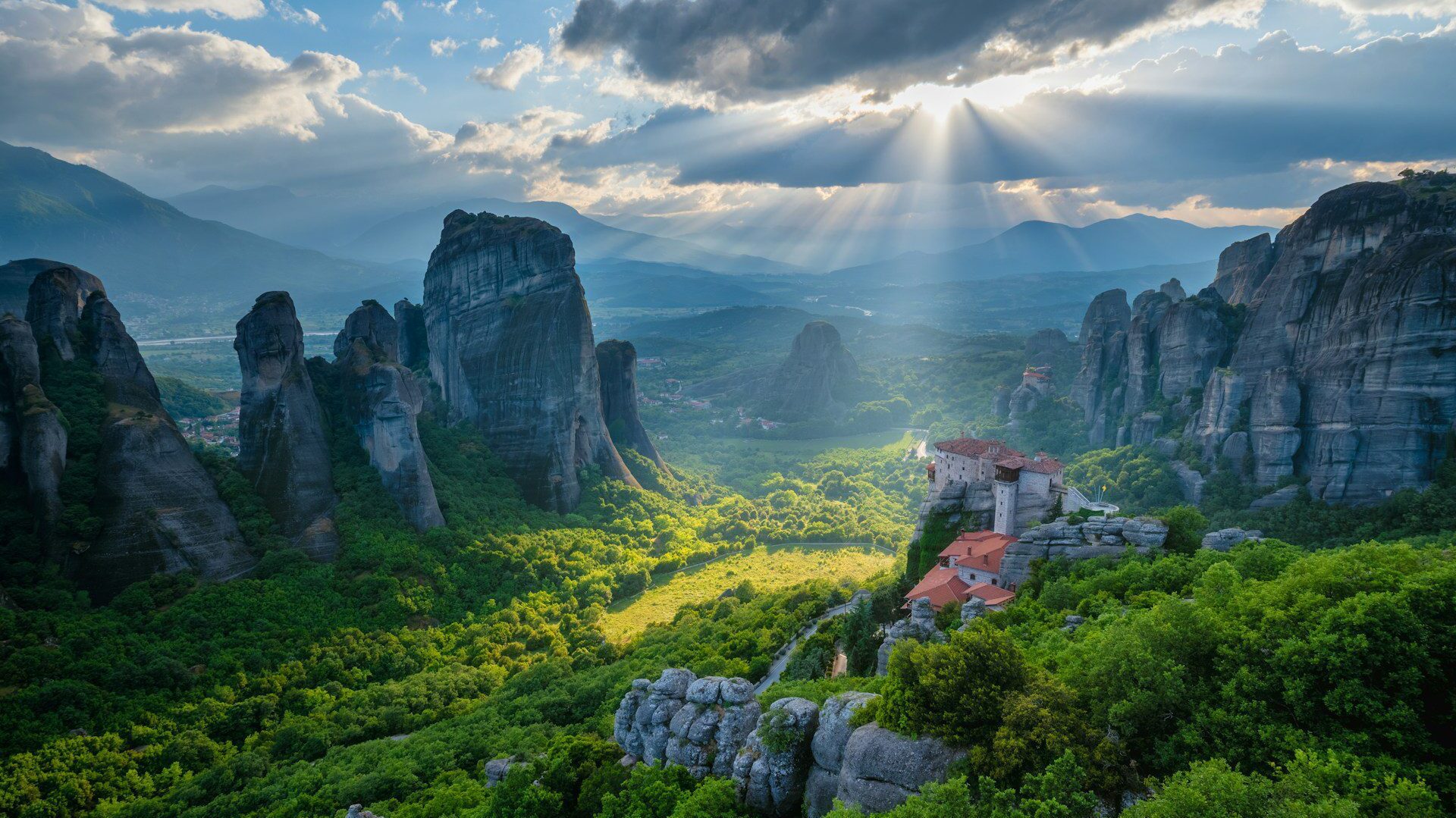 A clifftop monastery overlooking a green landscape with towering rocky mountains rising up all around