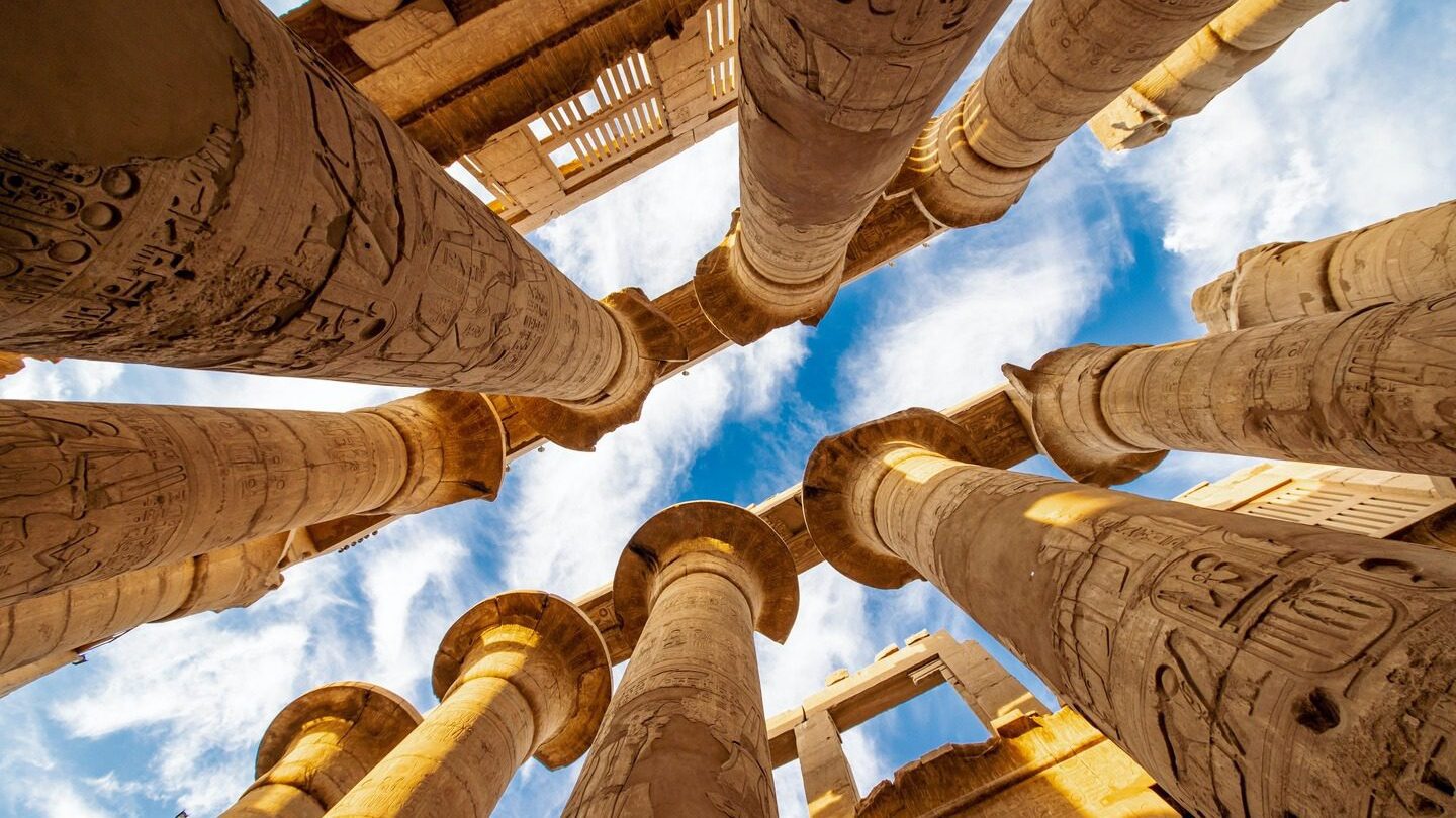 View from below of the towering columns at Karnak Temple in Egypt, showcasing intricate hieroglyphics against a blue sky with clouds during spring tours.