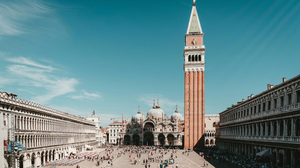A tall church tower overlooks Piazza San Marco with a domed building in the background in Venice.