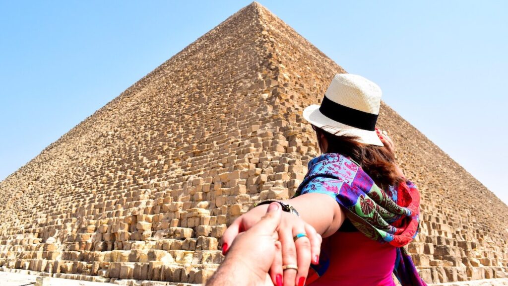 Woman in a hat, dressed for a trip to Egypt, being led by the hand towards the great pyramid of Giza.
