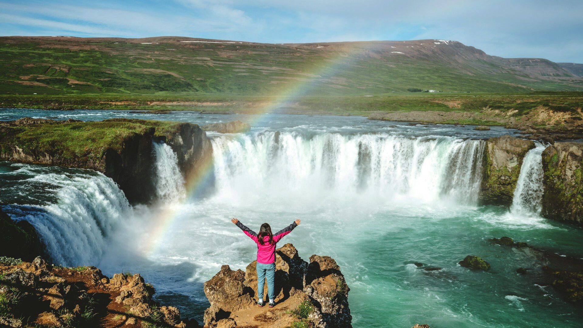 Woman standing on rocks overlooking a waterfall in Iceland with a rainbow over it 
