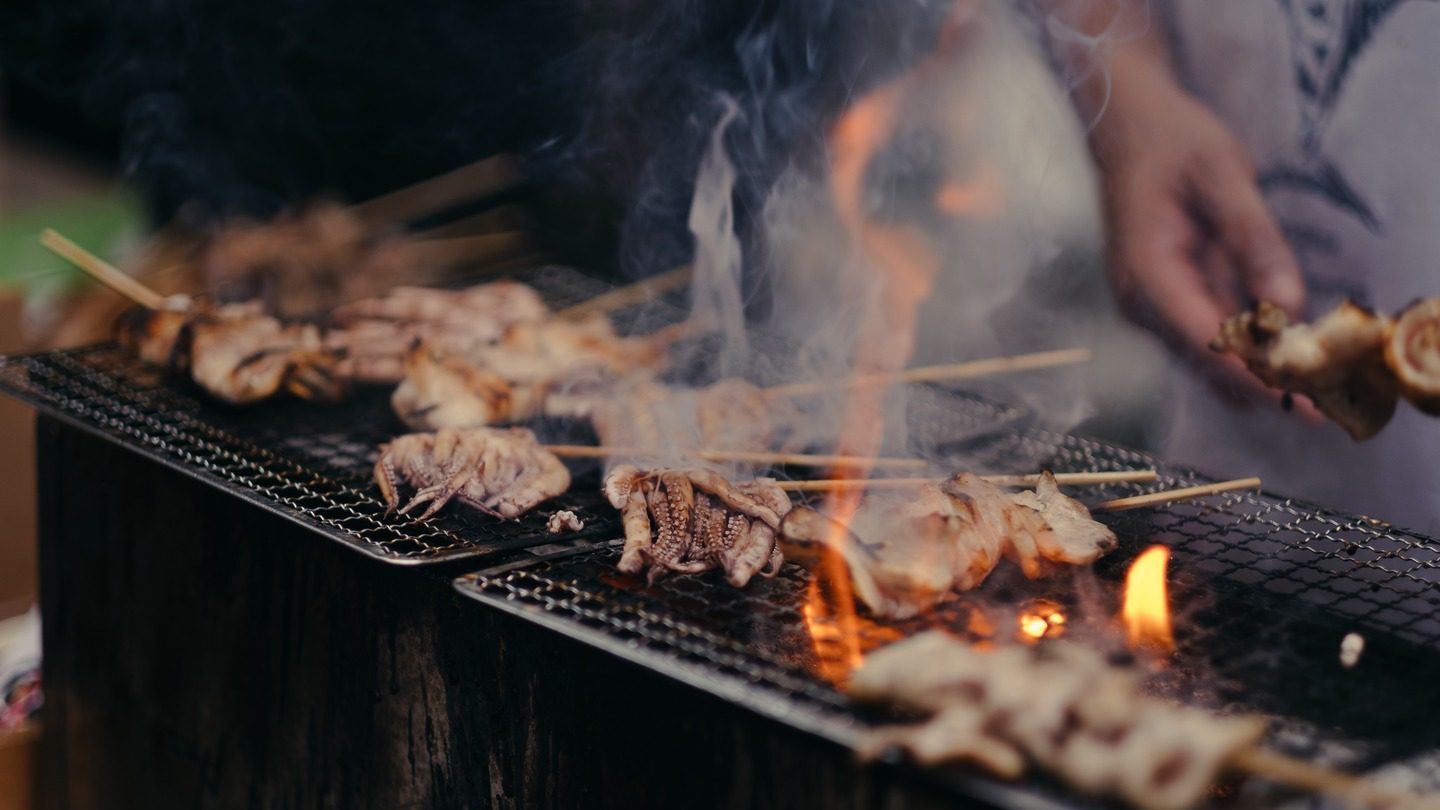 Skewered meat cooking on a flaming grill during a cultural holiday, with visible smoke and a person in the background adjusting the skewers.