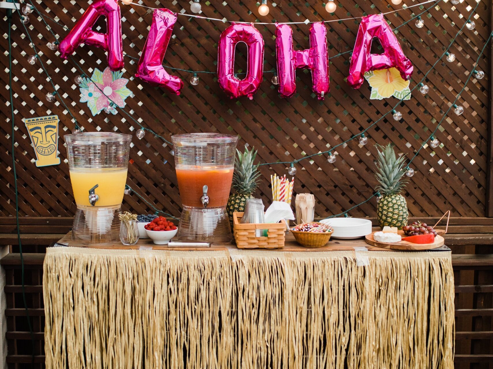 Outdoor party table decorated with a Hawaiian phrase "aloha" balloon sign, grass skirt trim, and drinks under string lights.
