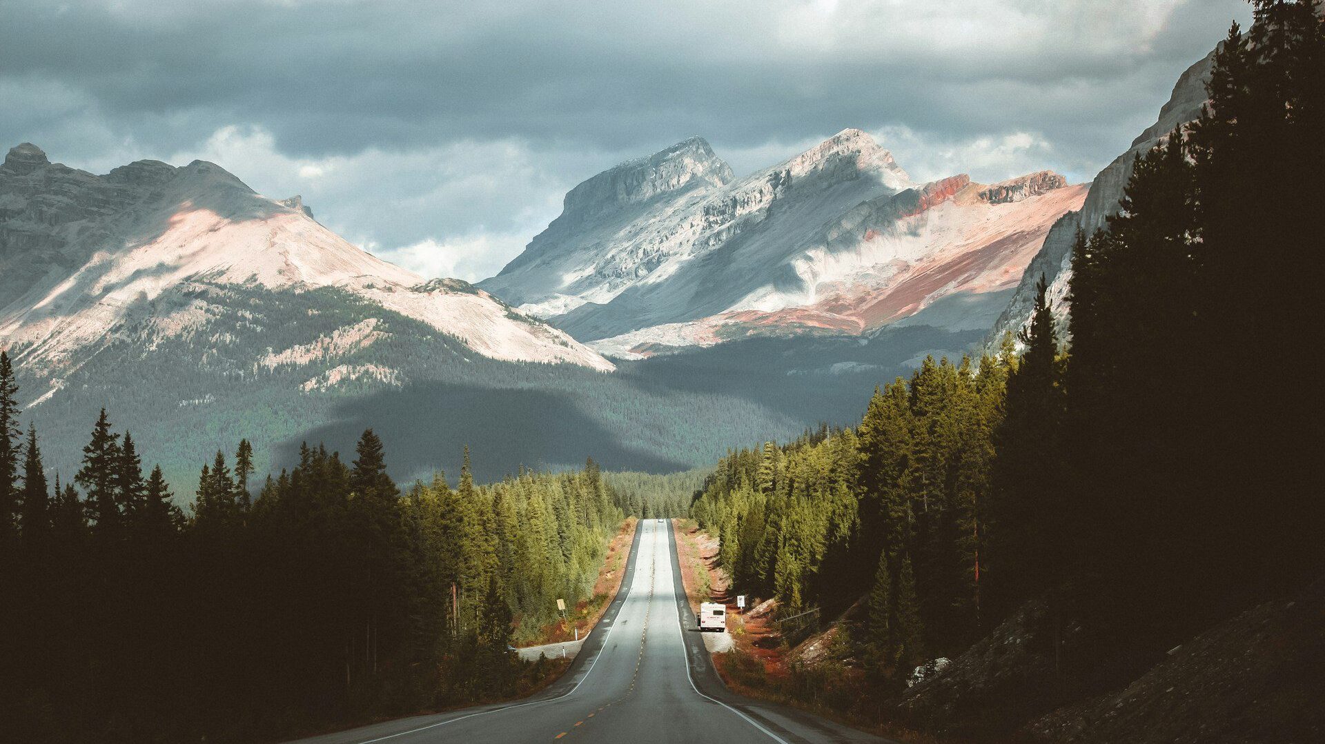 A straight road leading towards a snow-capped mountain, flanked by dense pine forests under a cloudy sky, perfect for cycling breaks.