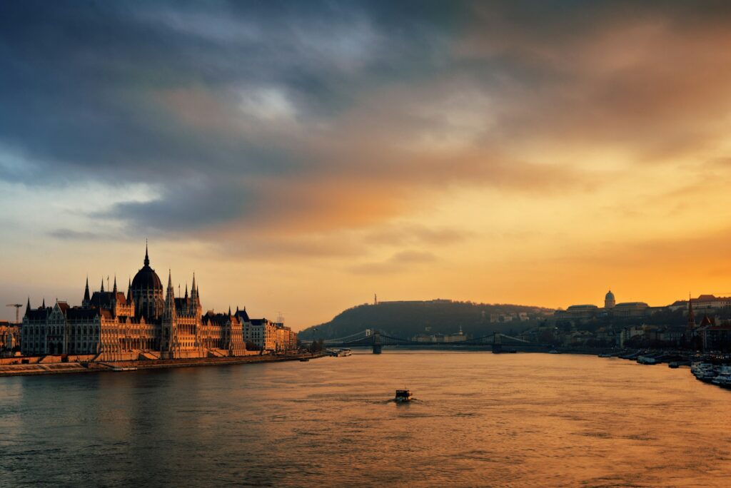 Looking down the Danube as it flows past the Hungarian houses of parliament in Budapest. Sky is cloudy, glowing orange at sunset.