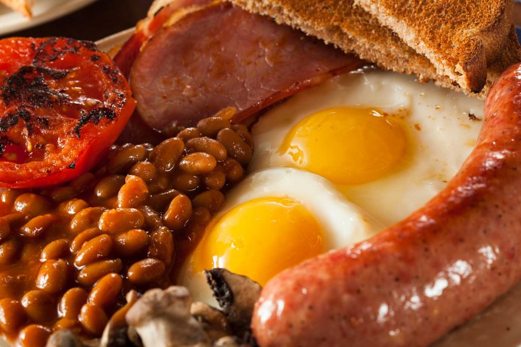 Close up of a full English breakfast with fried eggs, baked beans, sausages, toast, bacon and a grilled tomato.