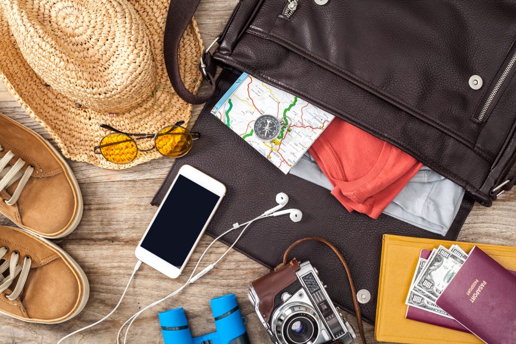 A satchel bag with travel items spilling out including an iphone with headphone, a camera, passport with cash, sunglasses, map, clothing, shoes and a straw hat