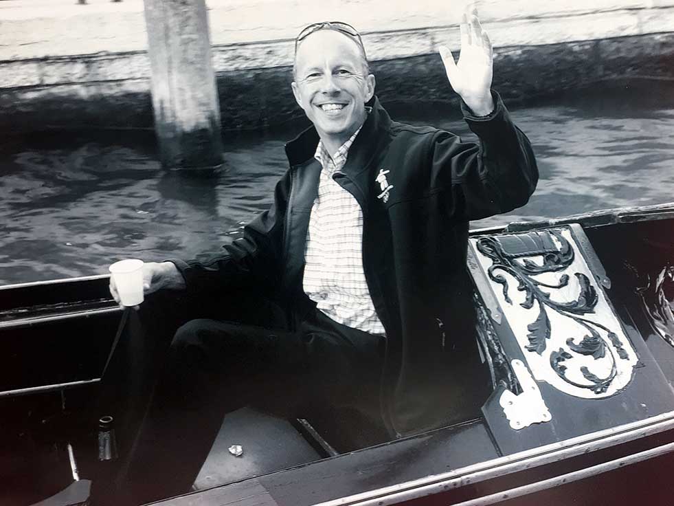 A man in a gondola waving to the camera during his Europe itinerary.