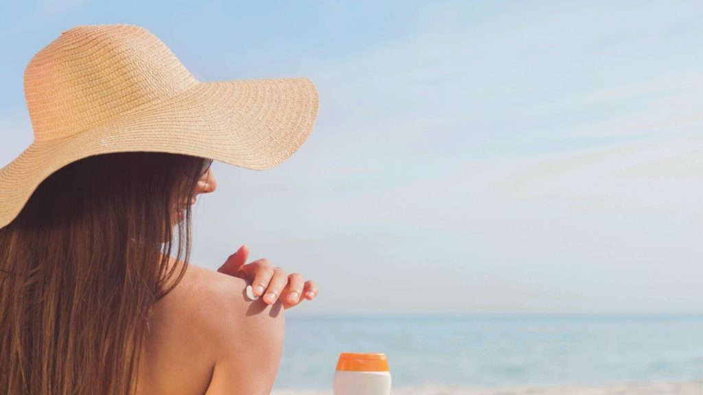 A woman wearing a hat on the beach with a bottle of sunscreen.