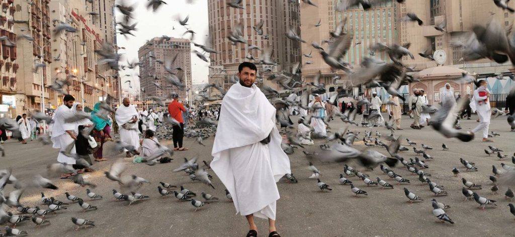 A man in traditional white clothing, typical of what to wear in the Middle East, stands among flying pigeons in a bustling public square.