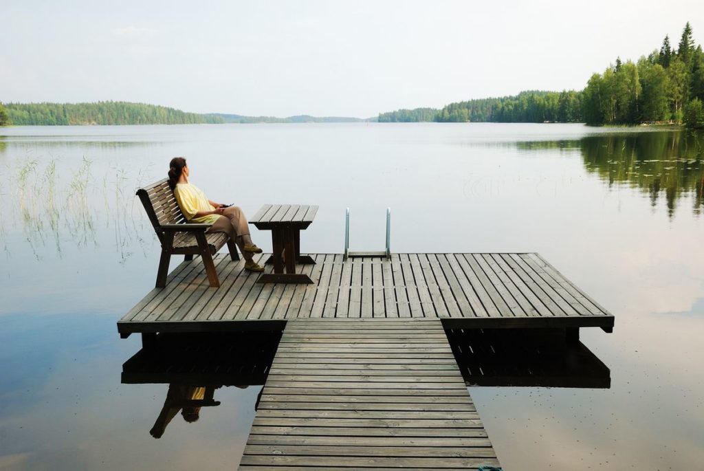 Person sitting on a bench at the end of a wooden pier, looking out over a calm lake surrounded by trees, dressed in scandi-cool fashion.