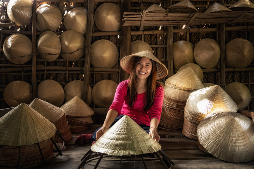 A smiling person holding a conical hat, surrounded by many similar hats in a workshop, showcasing what to pack for Vietnam.