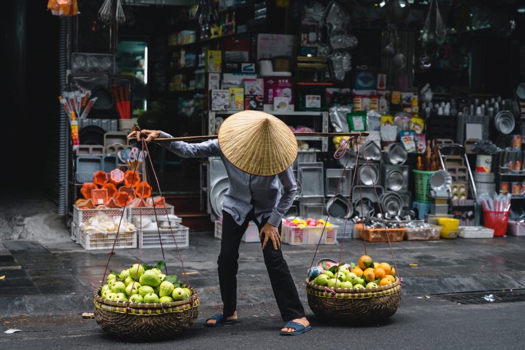 Vendor carrying fruit baskets on a pole across a traditional market street, showcasing what to pack for Vietnam.
