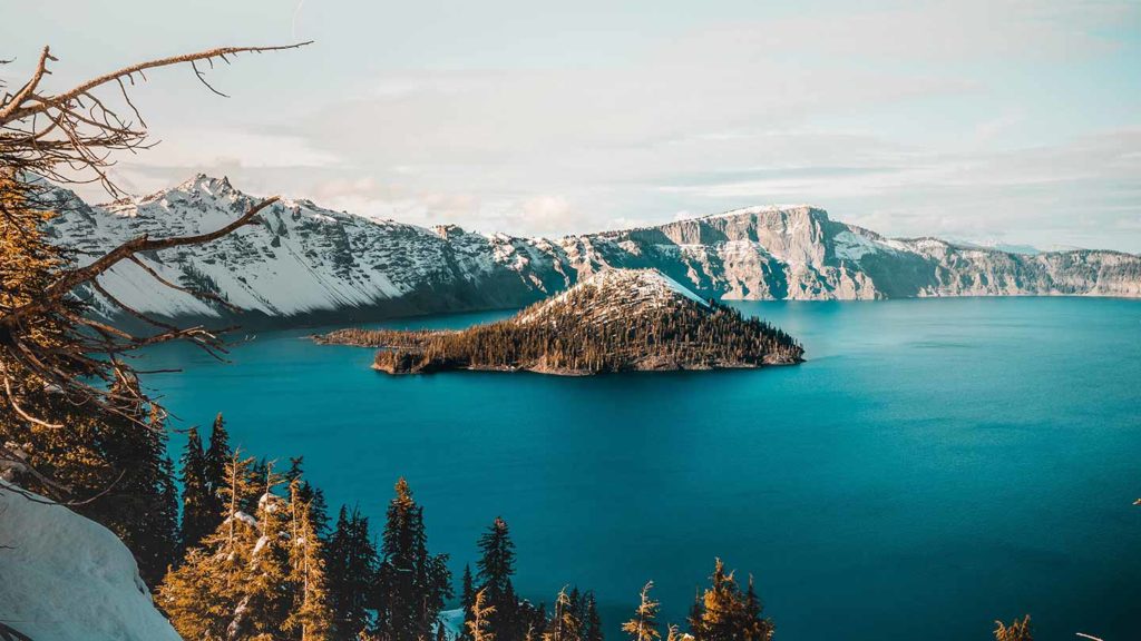 A serene view of Crater Lake with Wizard Island in the foreground, surrounded by snow-capped cliffs and conifer trees, highlighting one of the most beautiful American lakes.