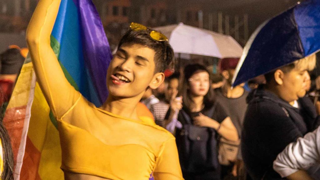 A person smiling and waving a rainbow flag at a pride 2021 event.