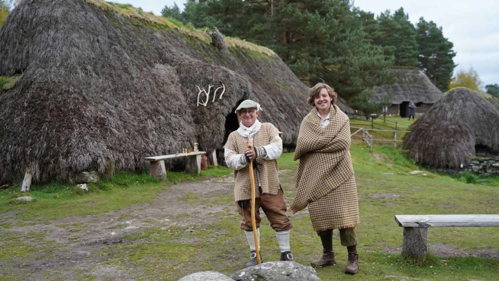 Two people in historical attire standing in front of thatched-roof buildings.