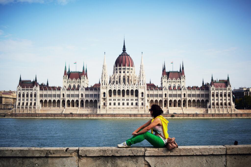 A female person sitting on a wall by the river, with Budapest's parliament building in the background.