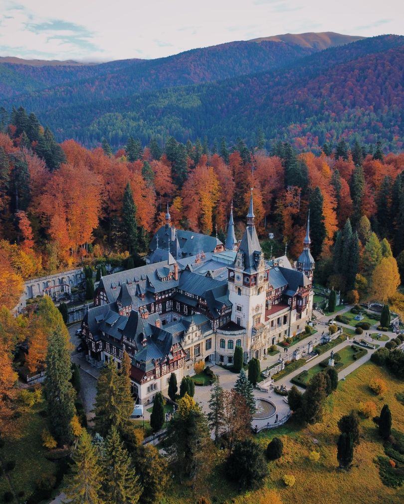Aerial view of a grand castle with pointed turrets surrounded by a dense autumn-colored forest under a soft evening light, ideal for family vacation destinations.