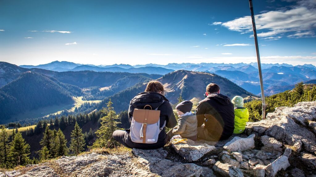 Three people on a multi-generational vacation sitting on a mountain peak, overlooking a vast range of forest-covered mountains under a clear blue sky.