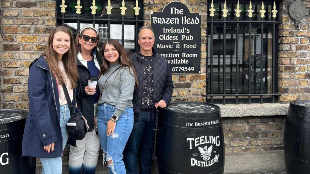 Four women smiling in front of the Brazen Head pub in Ireland, celebrating a daughter's graduation, standing beside barrels with the Teeling whiskey logo.