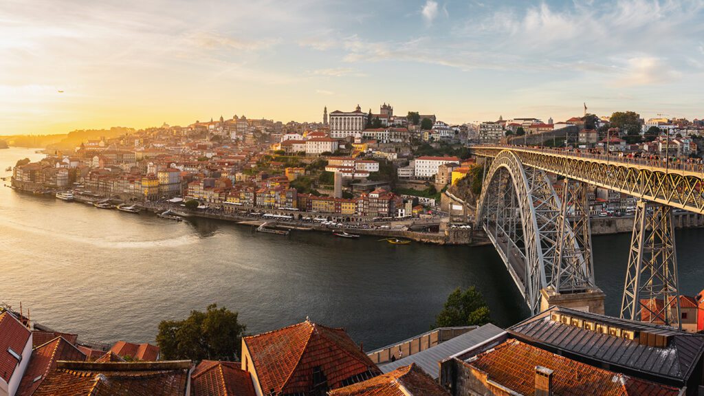 Panoramic view of Porto, Portugal, illustrating why travel to Portugal is a must, featuring the Douro River and Dom Luis I Bridge at sunset, with historic buildings spread across the hillside.