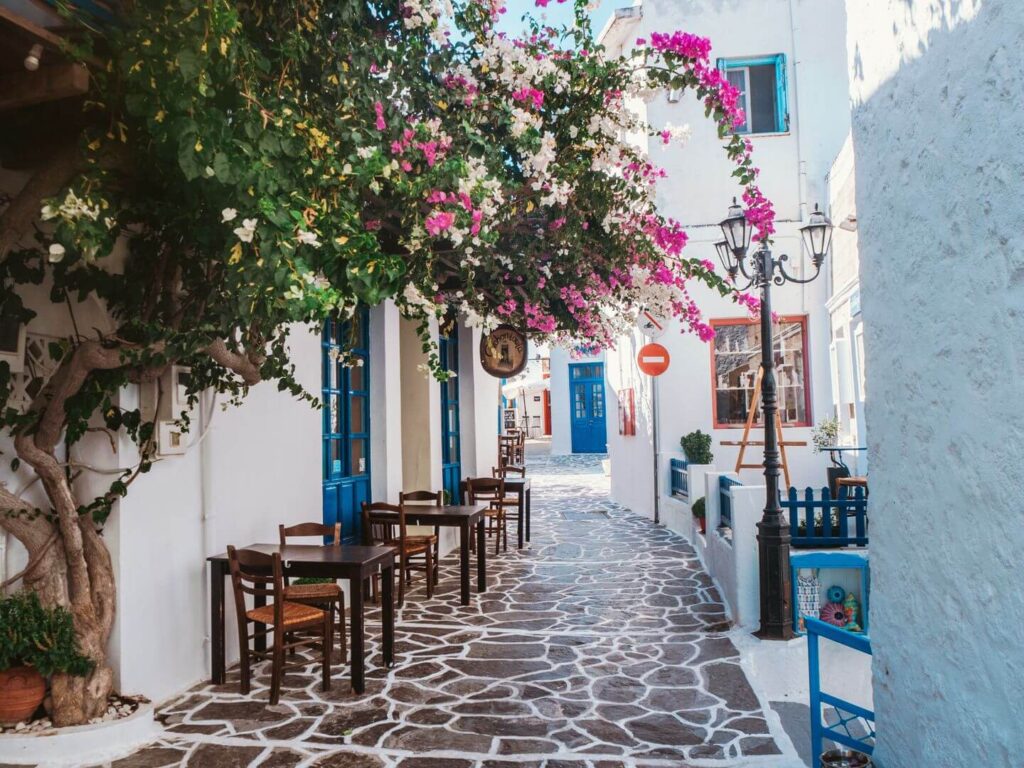 Charming alleyway in a Greek village with white-washed walls, cobblestone path, vibrant bougainvillea, and outdoor seating by blue doorways, ideal for tracing your heritage.