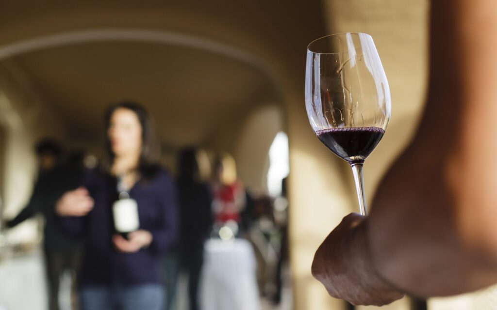 Close-up of a hand holding a glass of red wine, with blurred people socializing in the background at a local wine tasting event in Portugal.