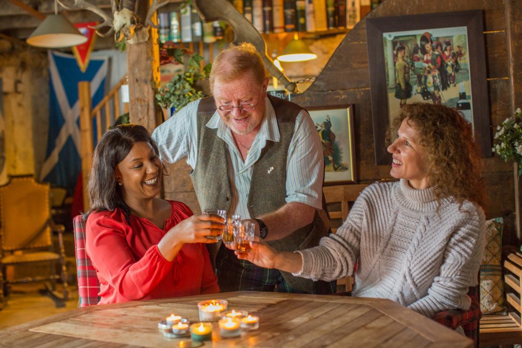 Three friends, a diverse group consisting of two women and a man, share a laugh over drinks at a rustic wooden table in a cozy, warmly-lit tavern during their 2022 travel rebound.