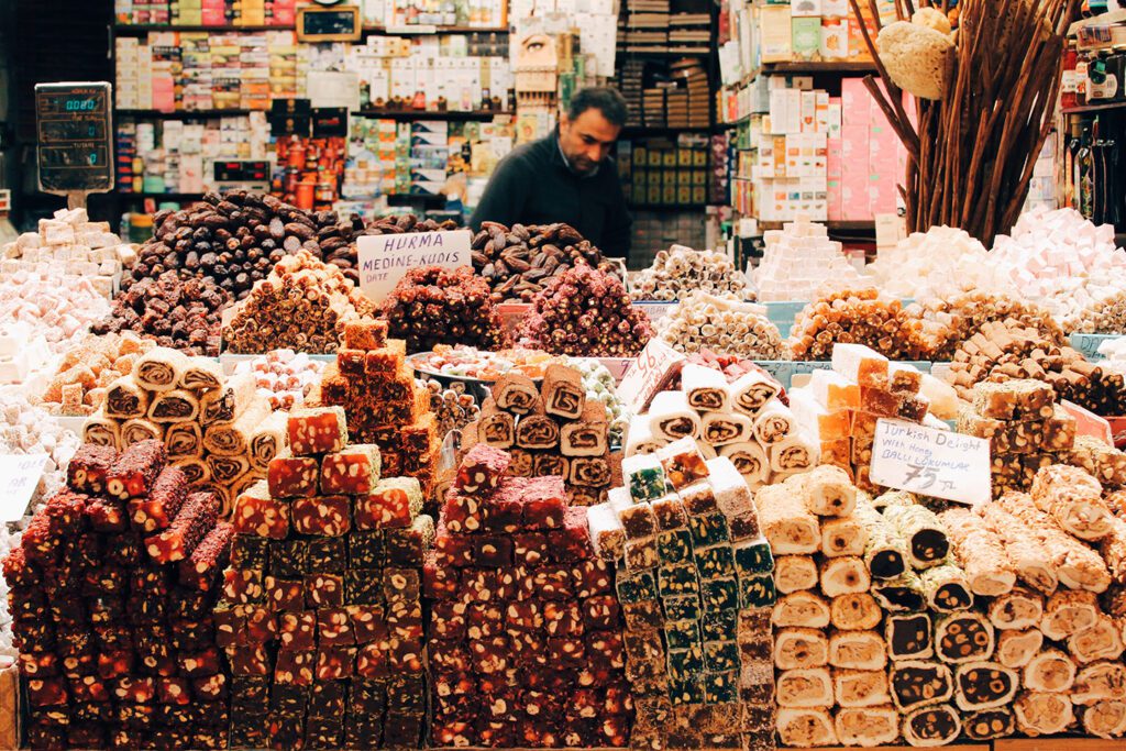 A vendor stands behind a colorful stall brimming with various types of Turkish delight and dried fruits at a bustling market in Turkey in 2022.