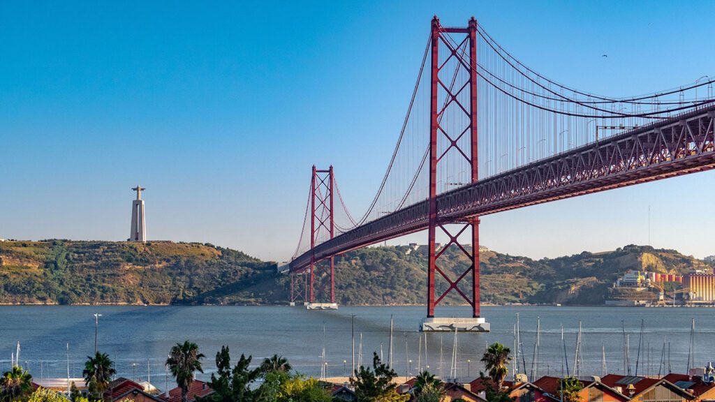 View of a red suspension bridge over a river, with a large statue on a hill in the background and rooftops in the foreground in Portugal.