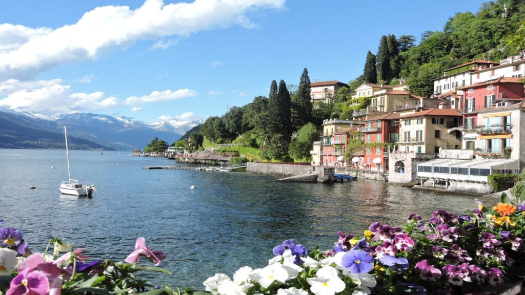 A scenic view of an Italian lakeside town with colorful buildings and boats, nestled against a backdrop of mountains and blue sky, framed by vibrant flowers in the foreground.