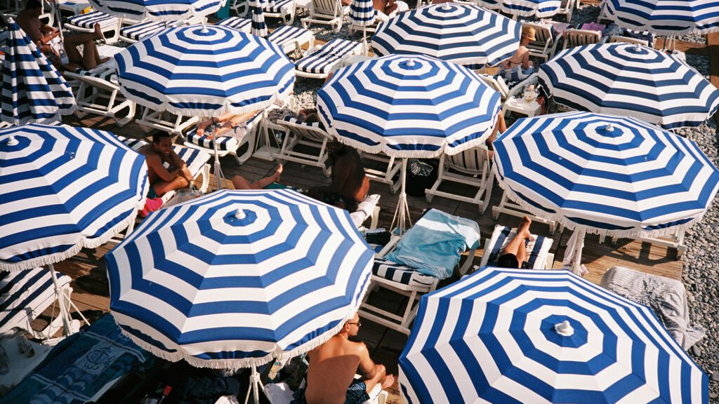 Aerial view of a crowded beach, one of the best beaches of Europe, with multiple blue and white striped umbrellas and sunbathers lounging on deck chairs.