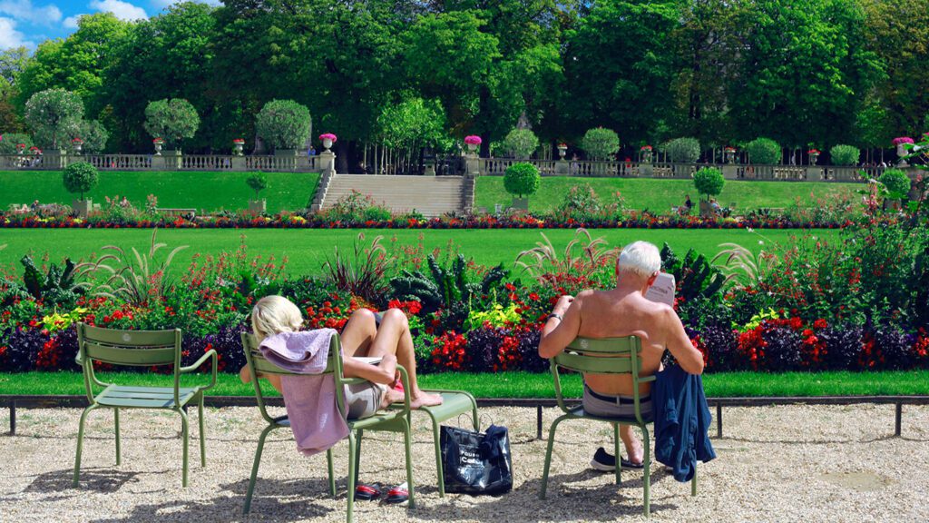 Two people sitting on park chairs facing a lush garden with vibrant flowers and trees under a clear summer Europe sky.