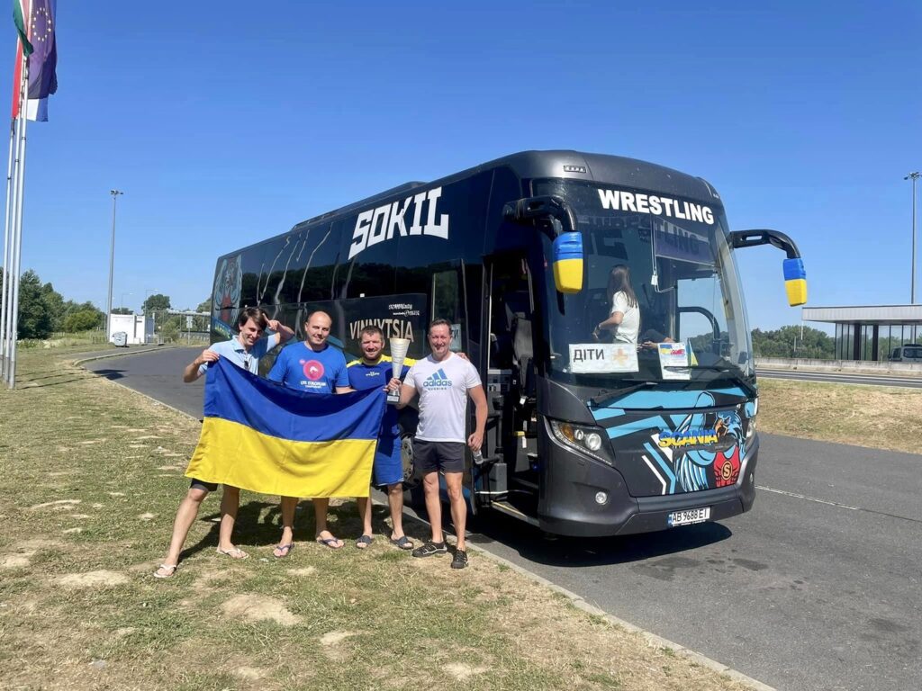 Four individuals posing with a Ukrainian flag in front of a "sokil" wrestling team bus on a sunny day, celebrating their Trafalgar Unlocked Award win.
