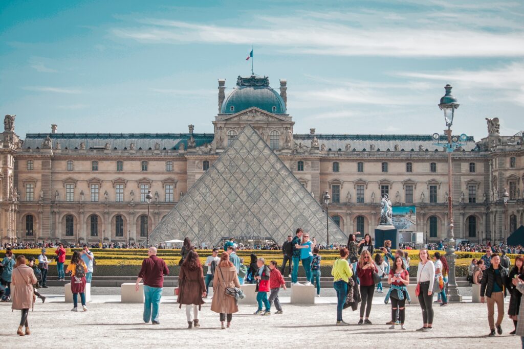 Tourists gather at the Louvre's courtyard, a highlight in any travel magazine, featuring the iconic glass pyramid and historical palace facade in Paris.
